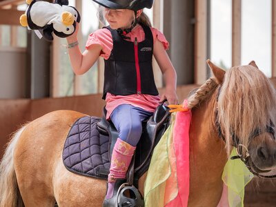 Kind mit Helm auf Pony sitzend, hält Plüschpinguin in der Hand in Reithalle