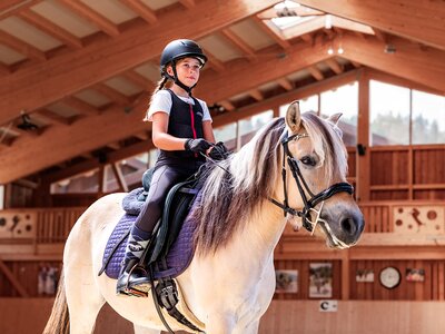 Mädchen mit Helm reitet Pferd in einer hellen Reithalle mit Holzbalken
