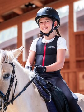 Mädchen mit Helm und Schutzweste reitet ein weißes Pferd in einer Reithalle