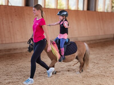 Junge Reitlehrerin führt Pony, auf dem ein Kind mit Helm und Schutzweste sitzt und die Arme ausstreckt