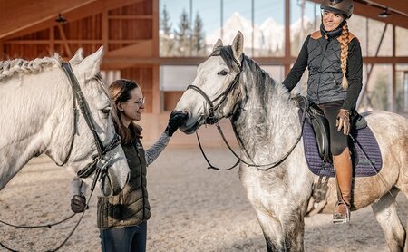 Frau berührt Pferd, während eine andere Frau auf einem zweiten Pferd in einer Reithalle sitzt